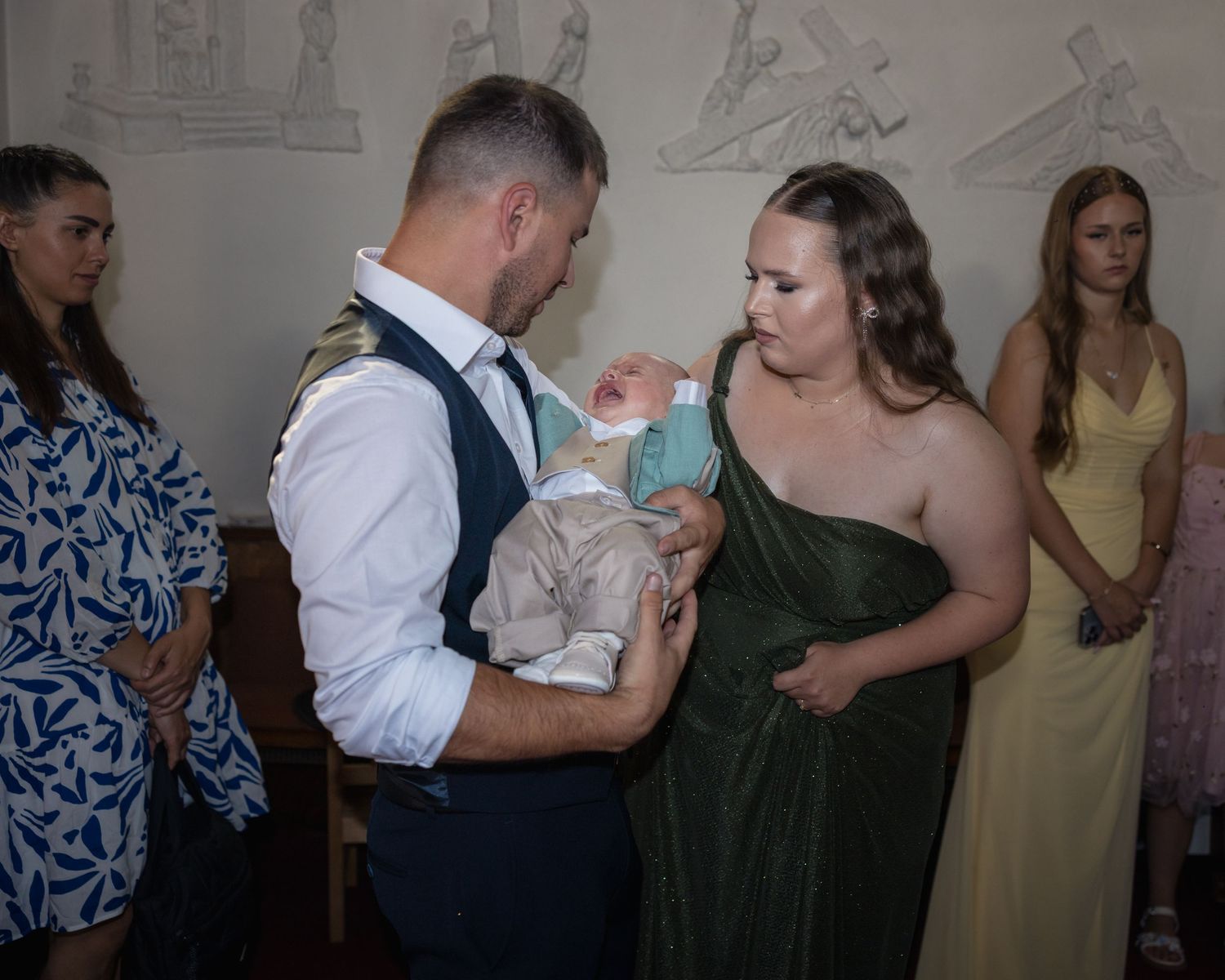 Ein Mann hält Theodor in der Kirche im Arm; eine Frau im grünen Kleid wendet sich dem Kind zu.