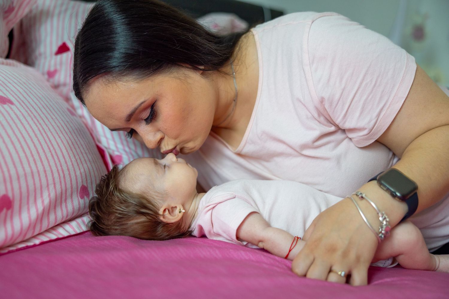 Eine Mutter küsst ihr Baby behutsam auf einem rosa Bett.