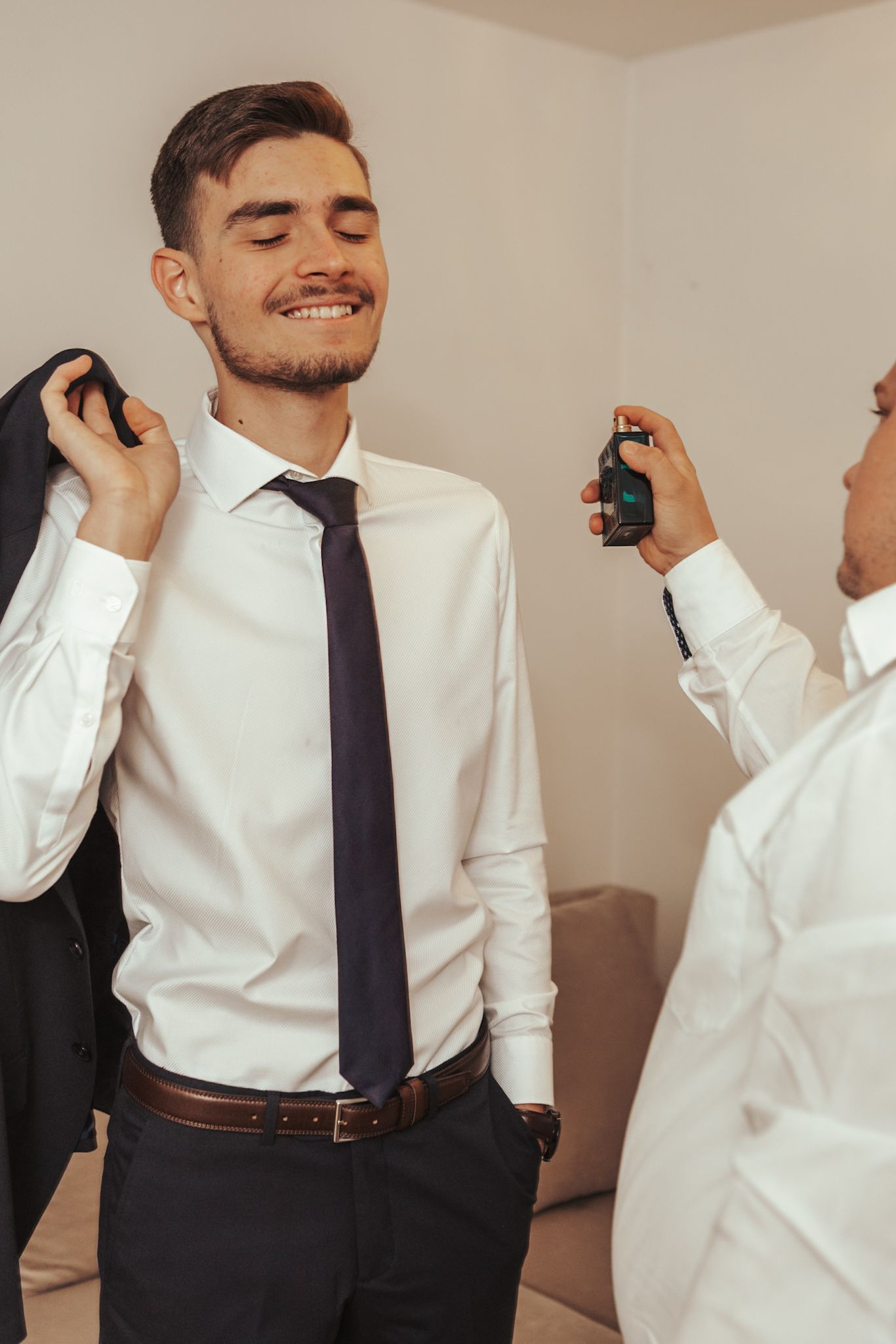 A smiling groom in a white shirt and tie gets cologne sprayed on him while holding his suit jacket before the wedding.