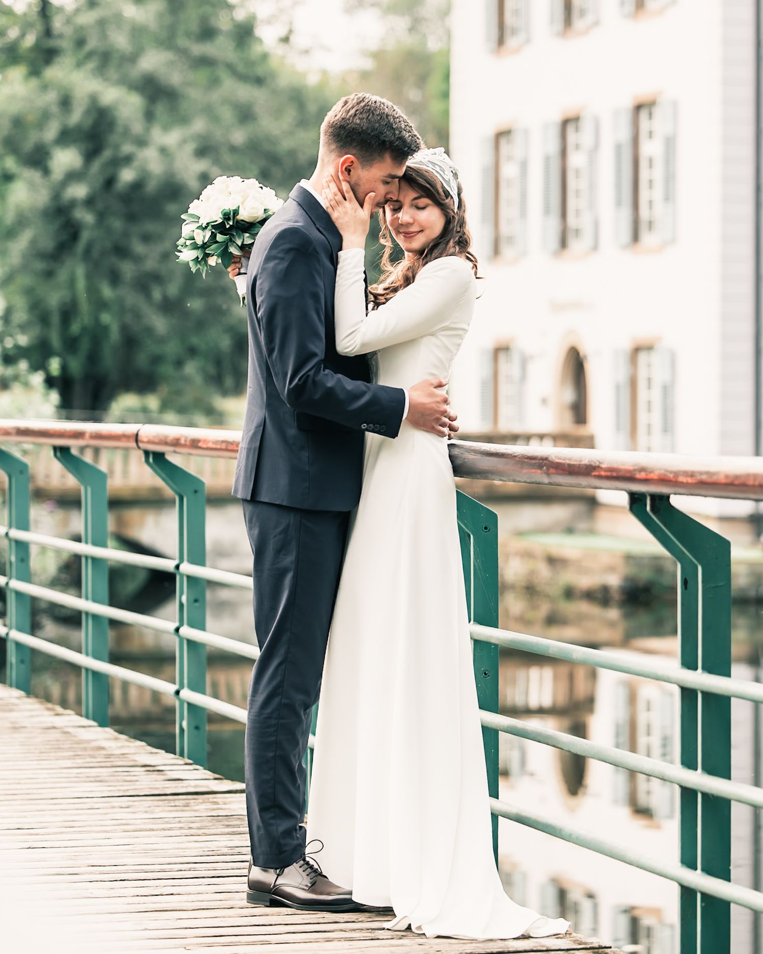 Bride and groom embracing on a bridge with white building in background, wedding day portrait outdoors.