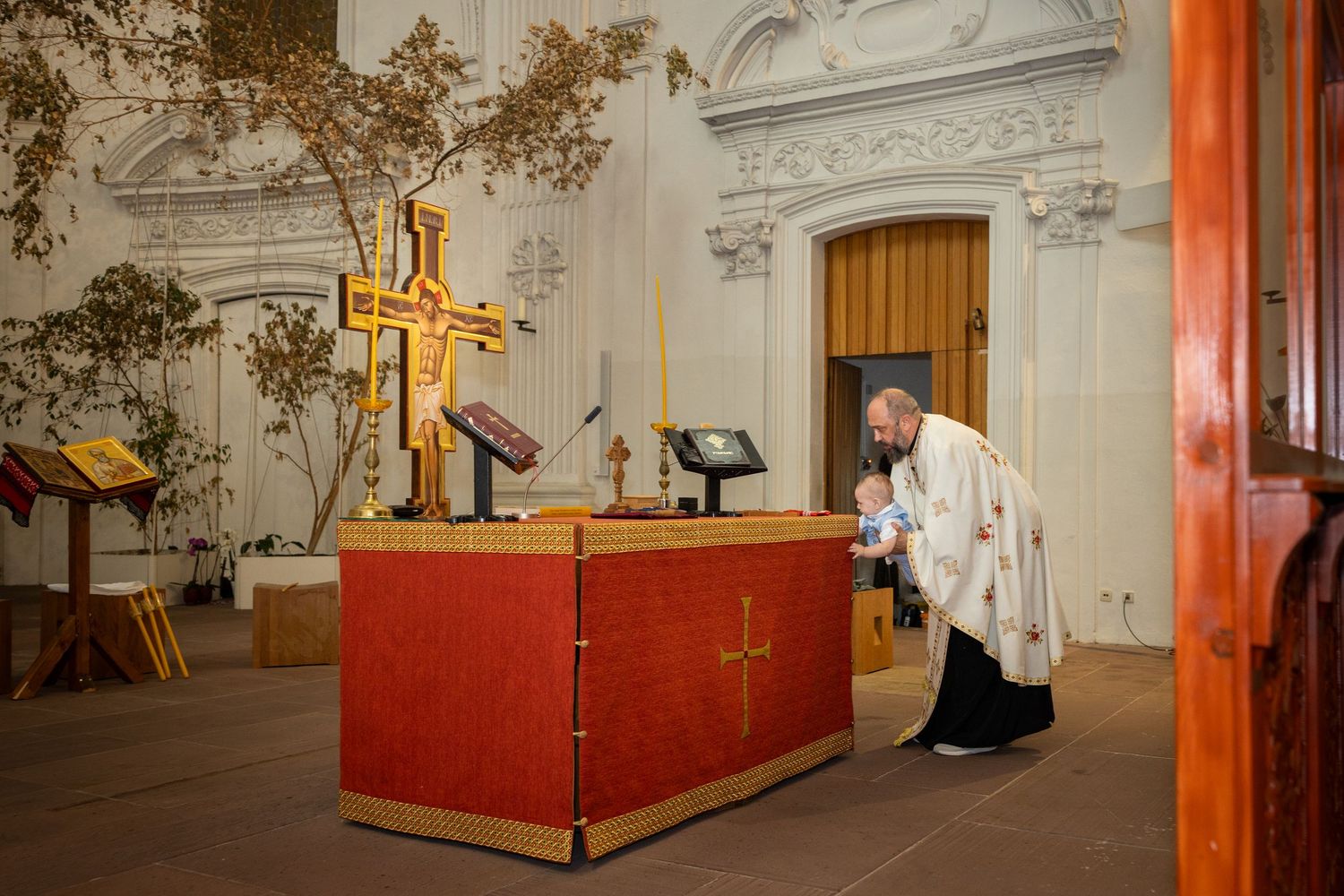 Der Priester trägt Aris an einen rot verkleideten Tisch im Kirchenraum; dahinter stehen Kreuz und Kerzen.