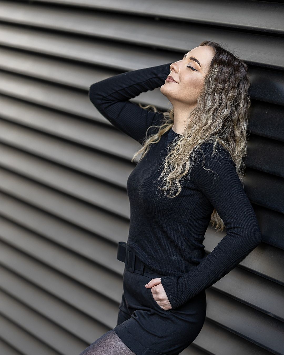 Woman in black dress posing confidently against striped wall with wavy blonde hair.