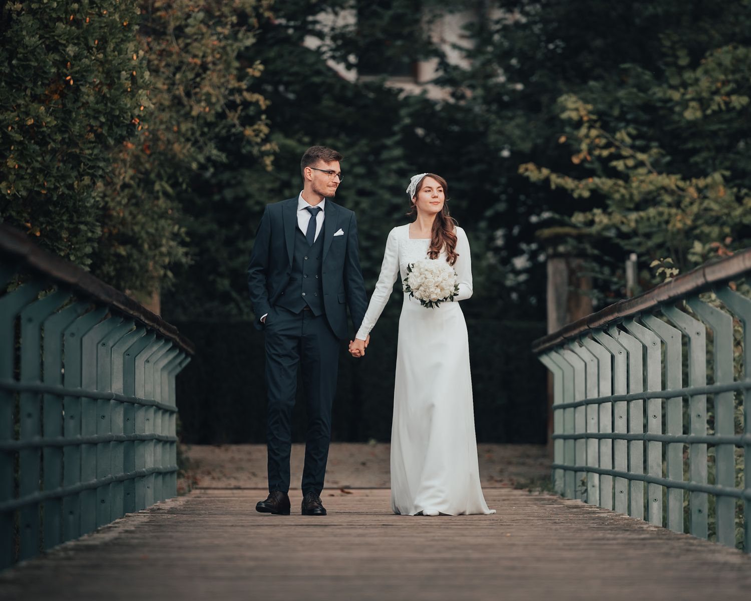 Bride and groom holding hands on a wooden bridge surrounded by lush green trees on their wedding day.