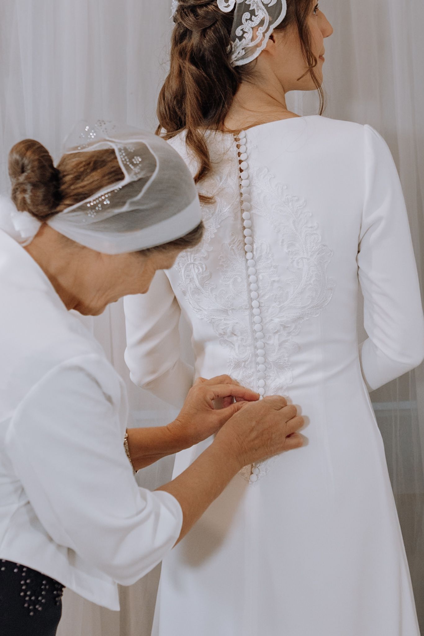 Bride in white wedding dress with button-back detail being helped by assistant holding decorative hat at wedding preparation.