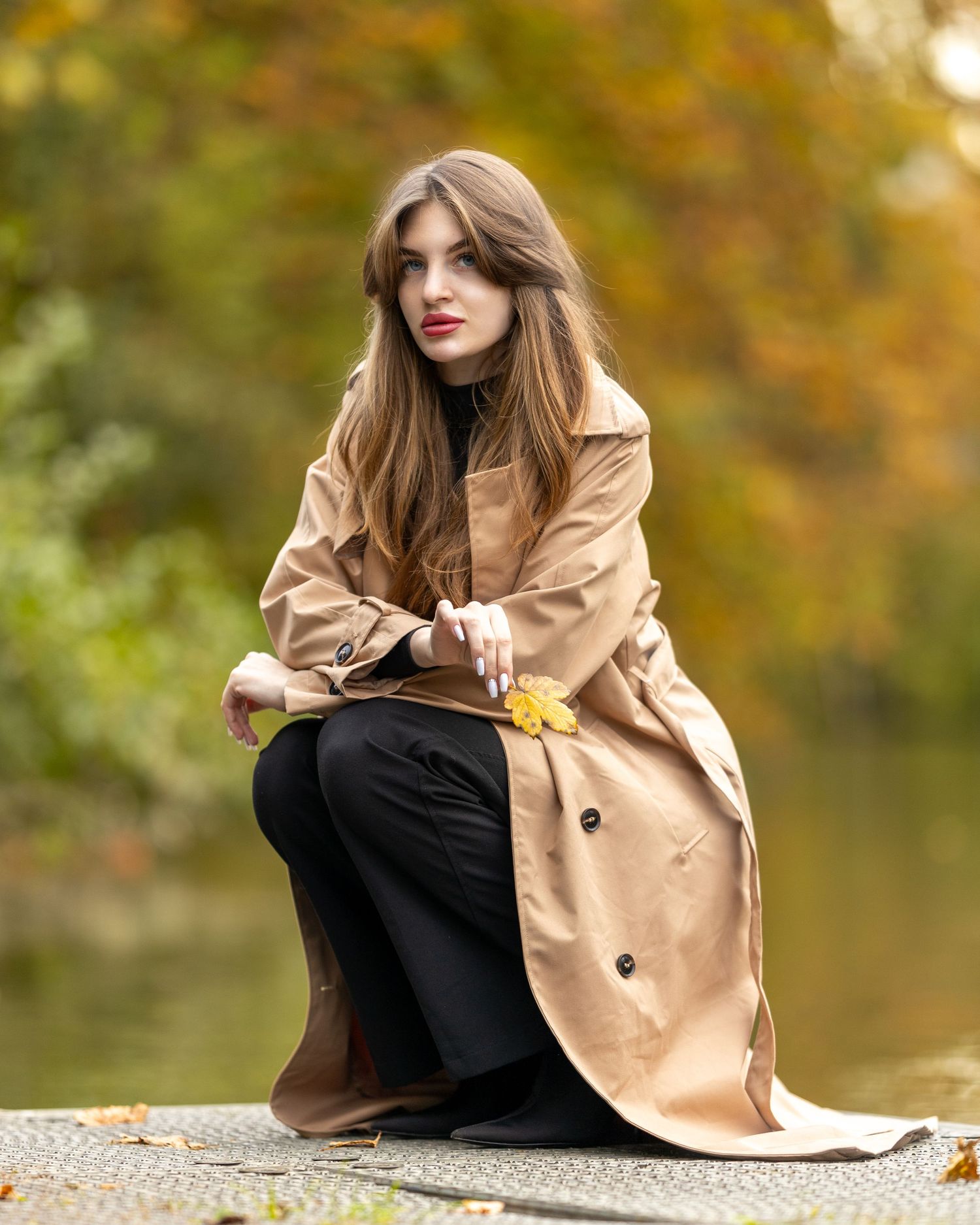 Frau im beigen Trenchcoat sitzt im herbstlichen Park vor buntem Laub.