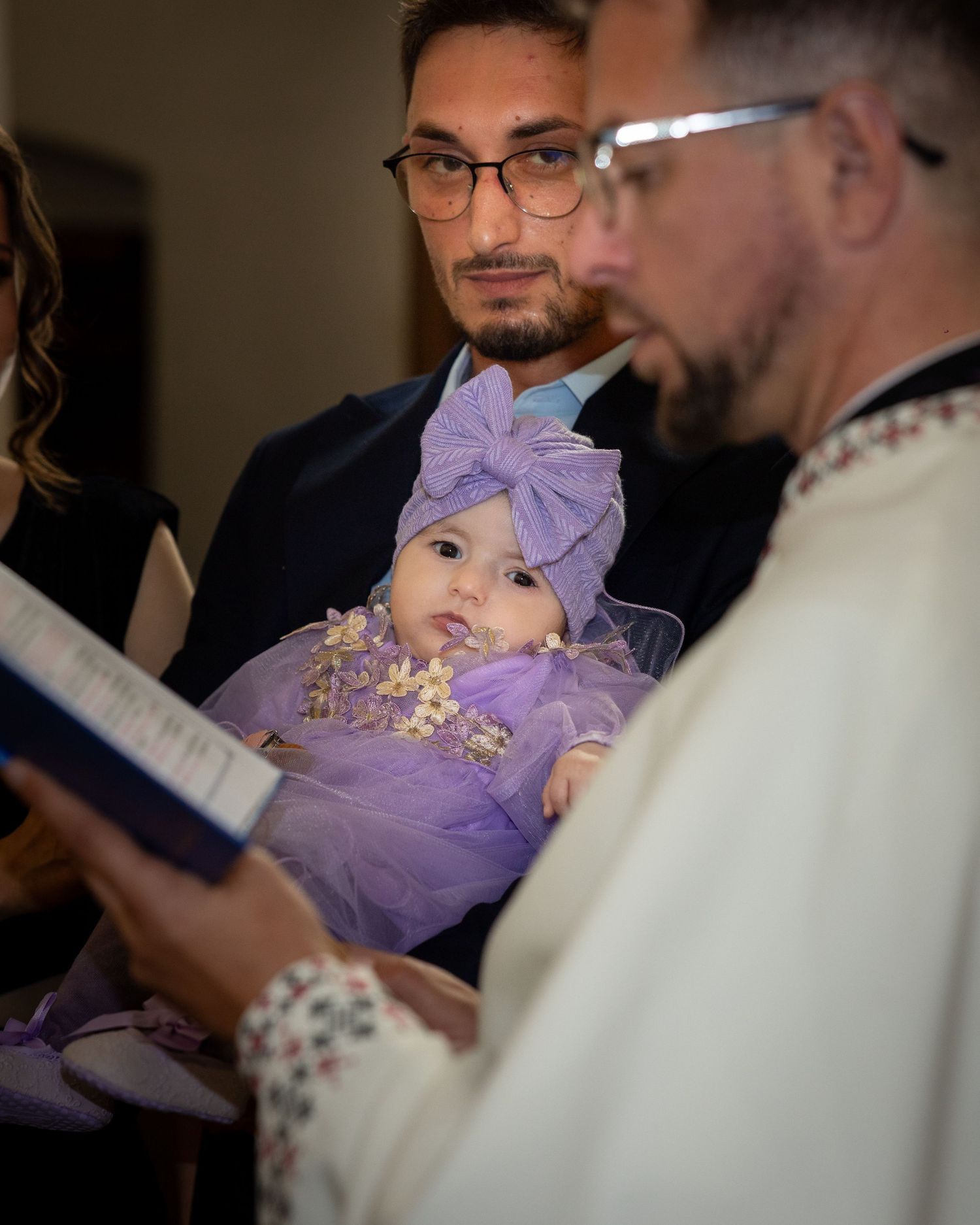 Baby girl in purple tulle dress and bow headband held during a church baptism ceremony.