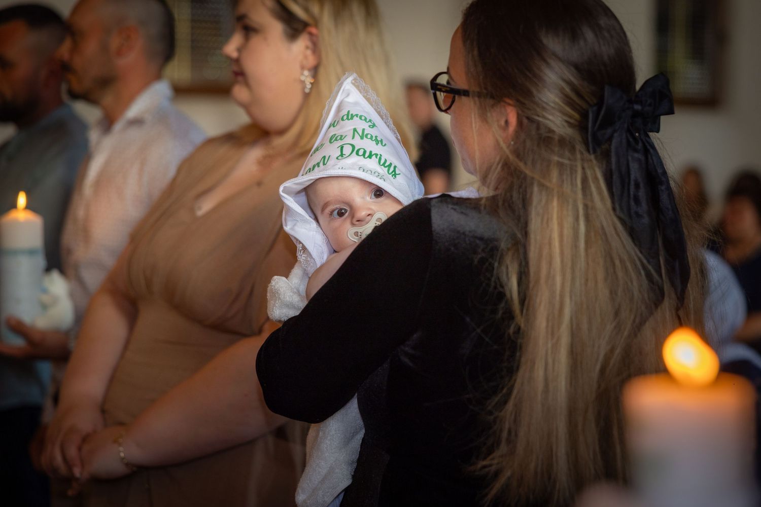 Baby in hooded baptism towel held by woman during christening ceremony with candles in church.