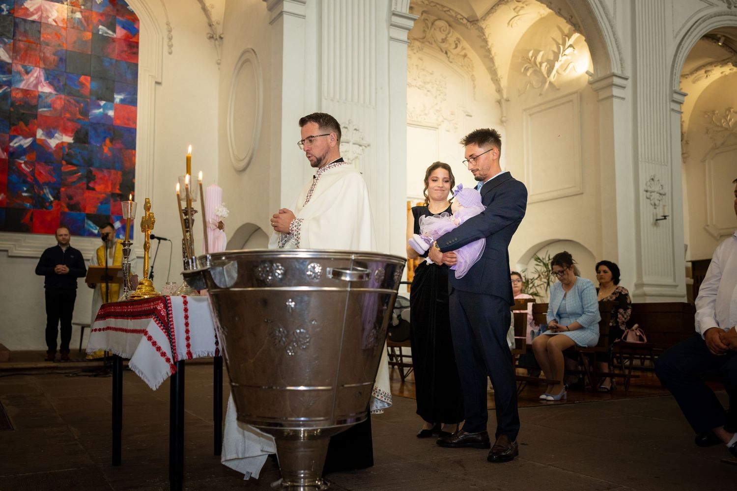 Priest performing a baptism ceremony at a church font with godparents standing beside a large decorative baptismal basin.