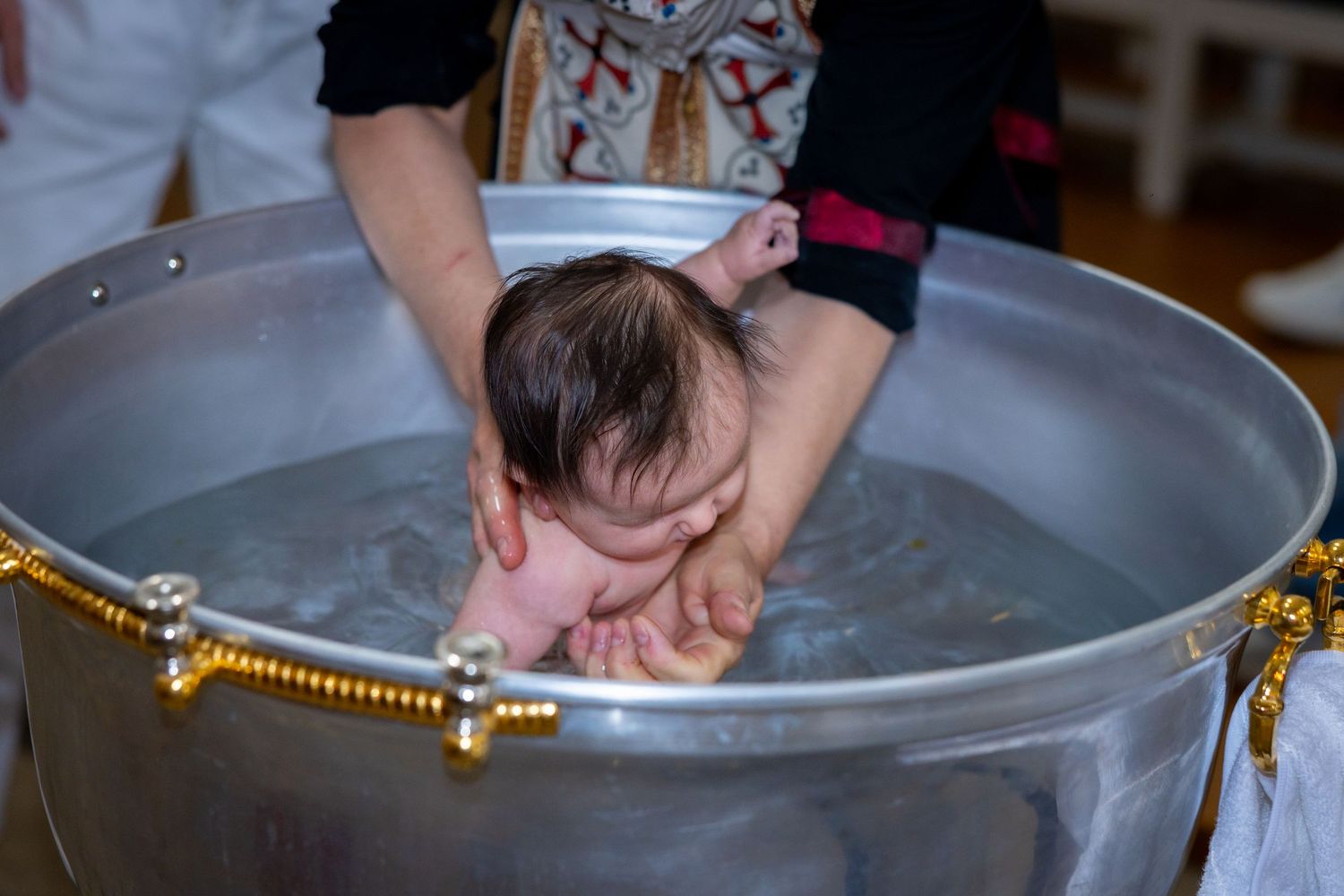 A newborn baby being baptized in a large silver baptismal font during a Christian christening ceremony.