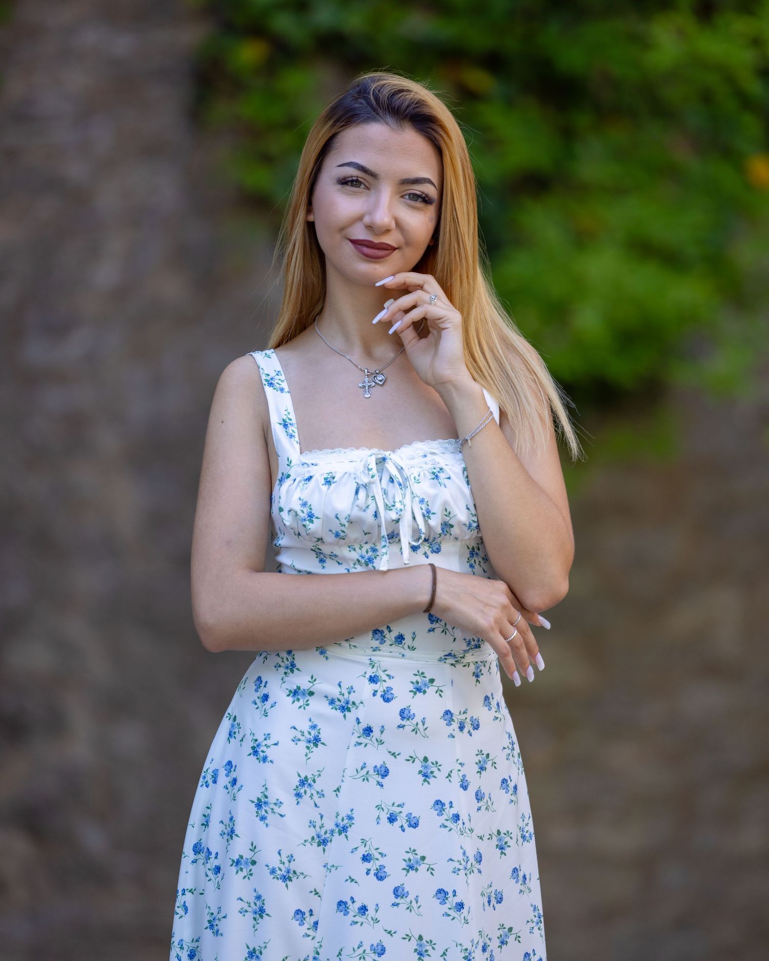 Young woman in white floral sundress smiling outdoors with green foliage background.