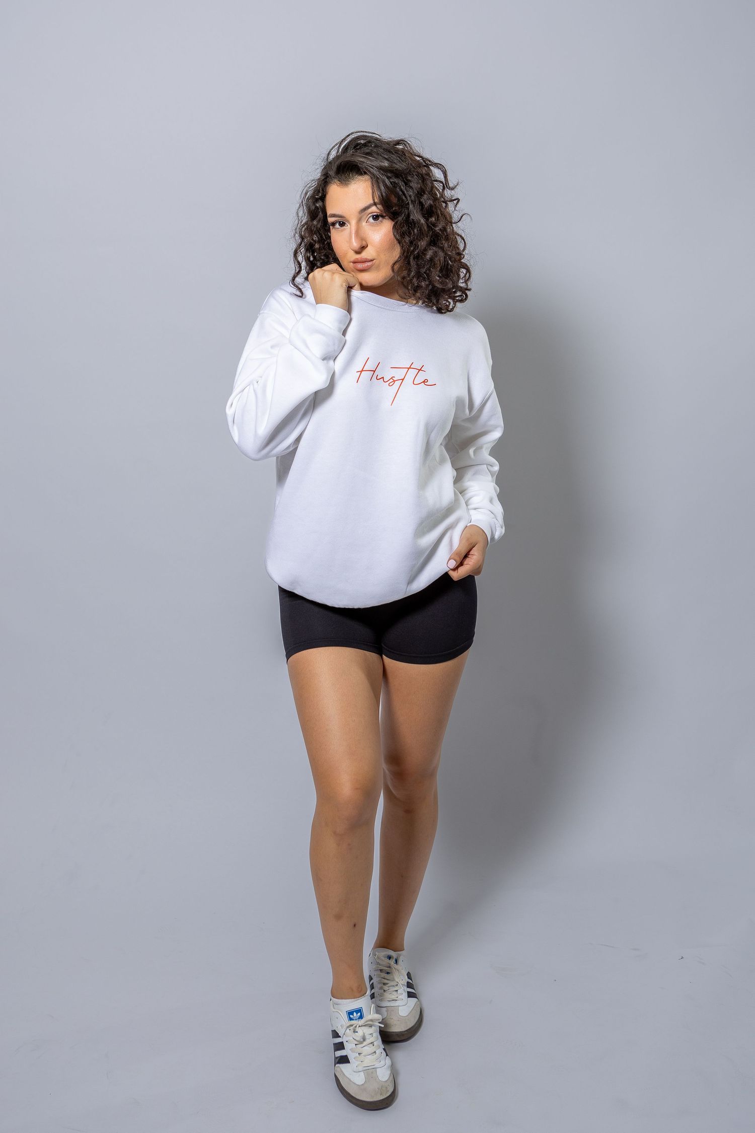 Young woman in white graphic sweatshirt, black shorts, and sneakers posing against gray background.
