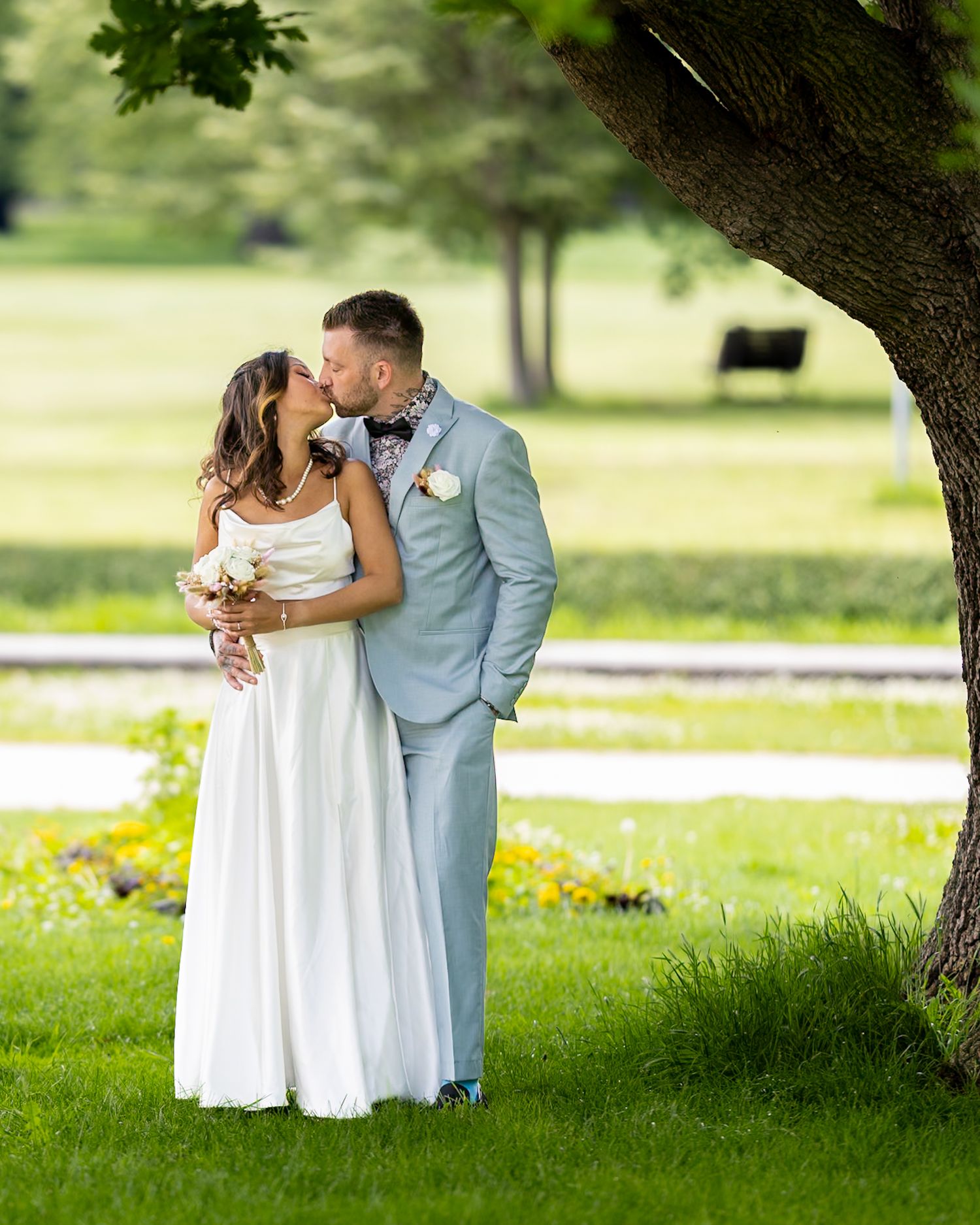 Bride and groom kissing under a tree in a sunny outdoor wedding setting with white dress and gray suit.