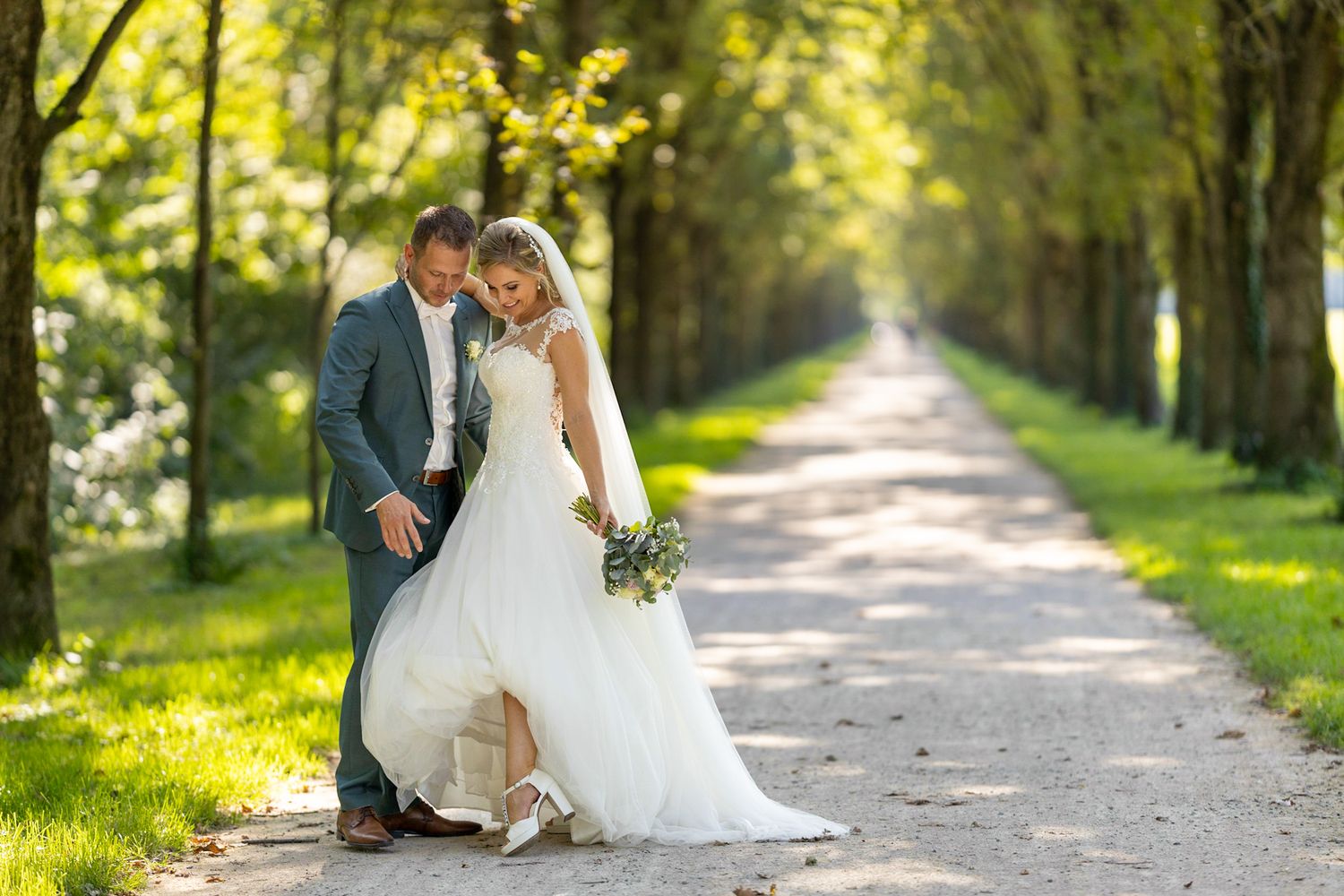 A bride and groom share a romantic moment on a tree-lined gravel path, bride in white gown holding a bouquet.