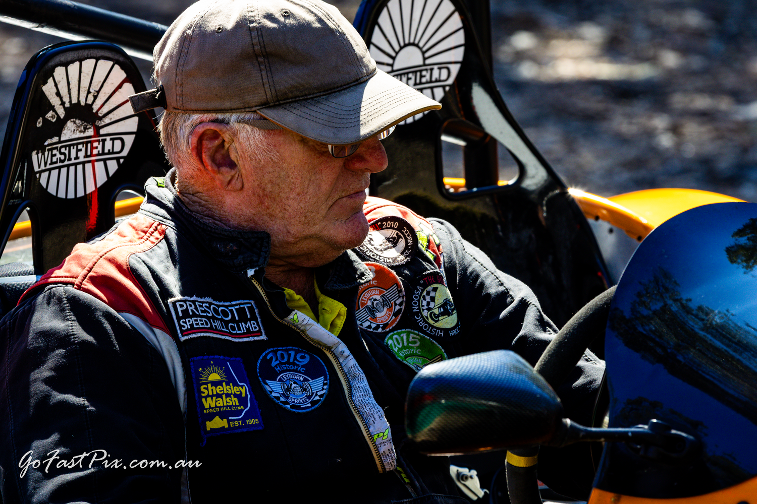 Experienced racing driver with patches on jacket sits beside yellow race car.