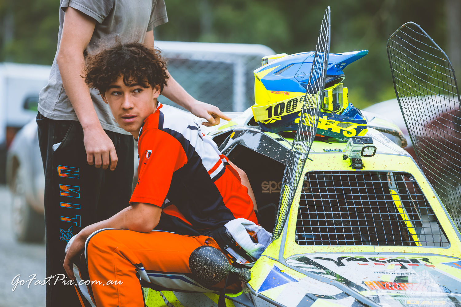 Young racing driver in orange suit sits on race car at motorsport event.