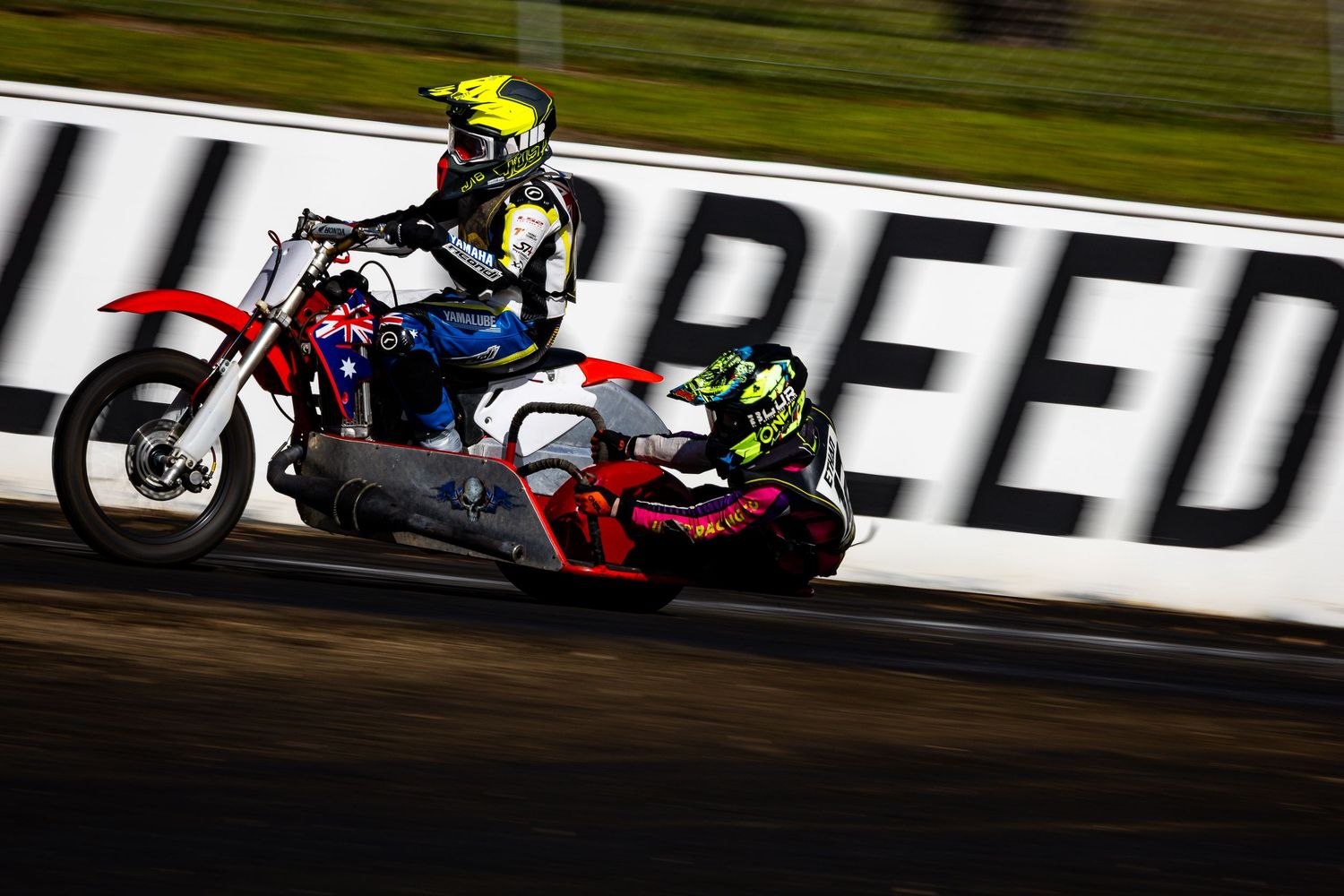 Two motorsport racers compete on a dirt track, one on a red motorcycle and another on a sidecar, with a speed banner behind.