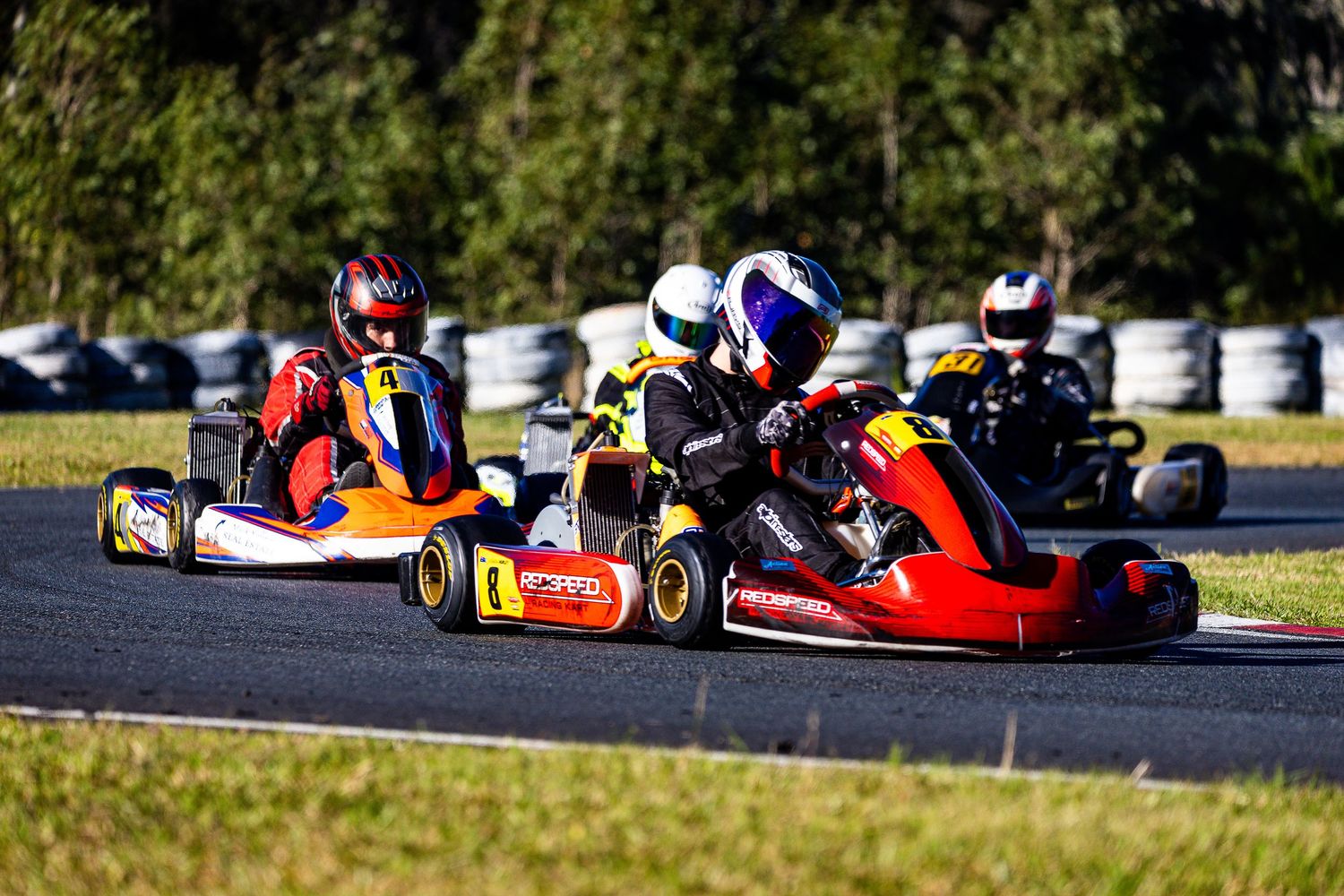 Go-kart racers compete on a winding outdoor track surrounded by trees during a sunny motorsport event.