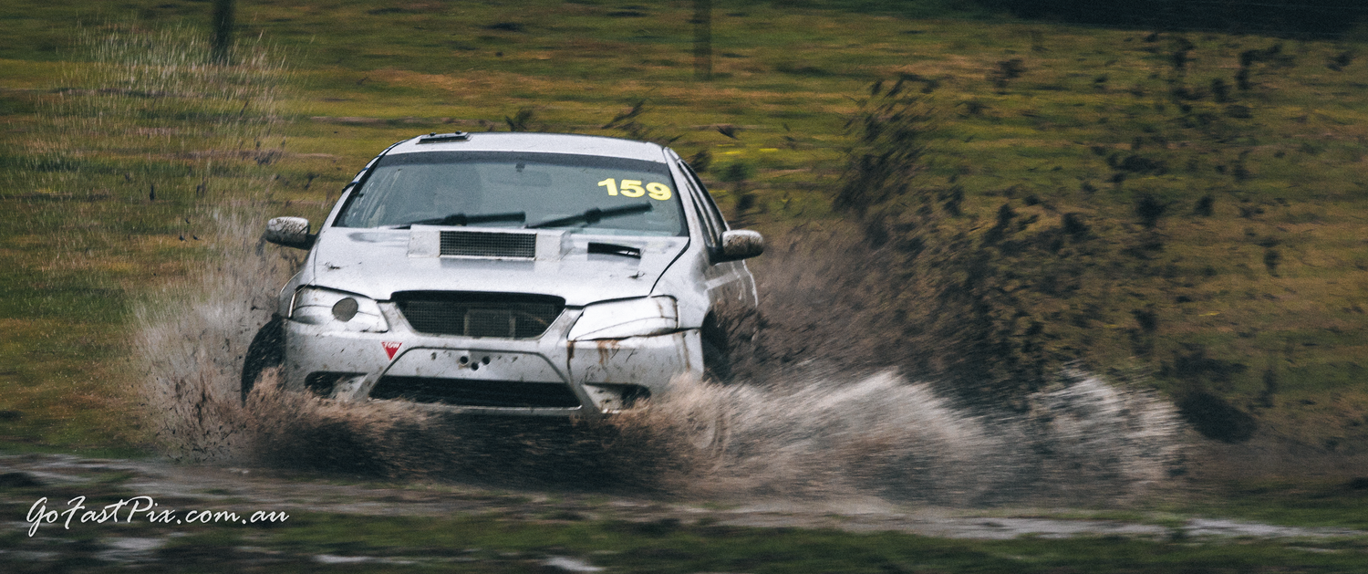 Silver rally car number 15 powers through muddy puddle on rally stage.