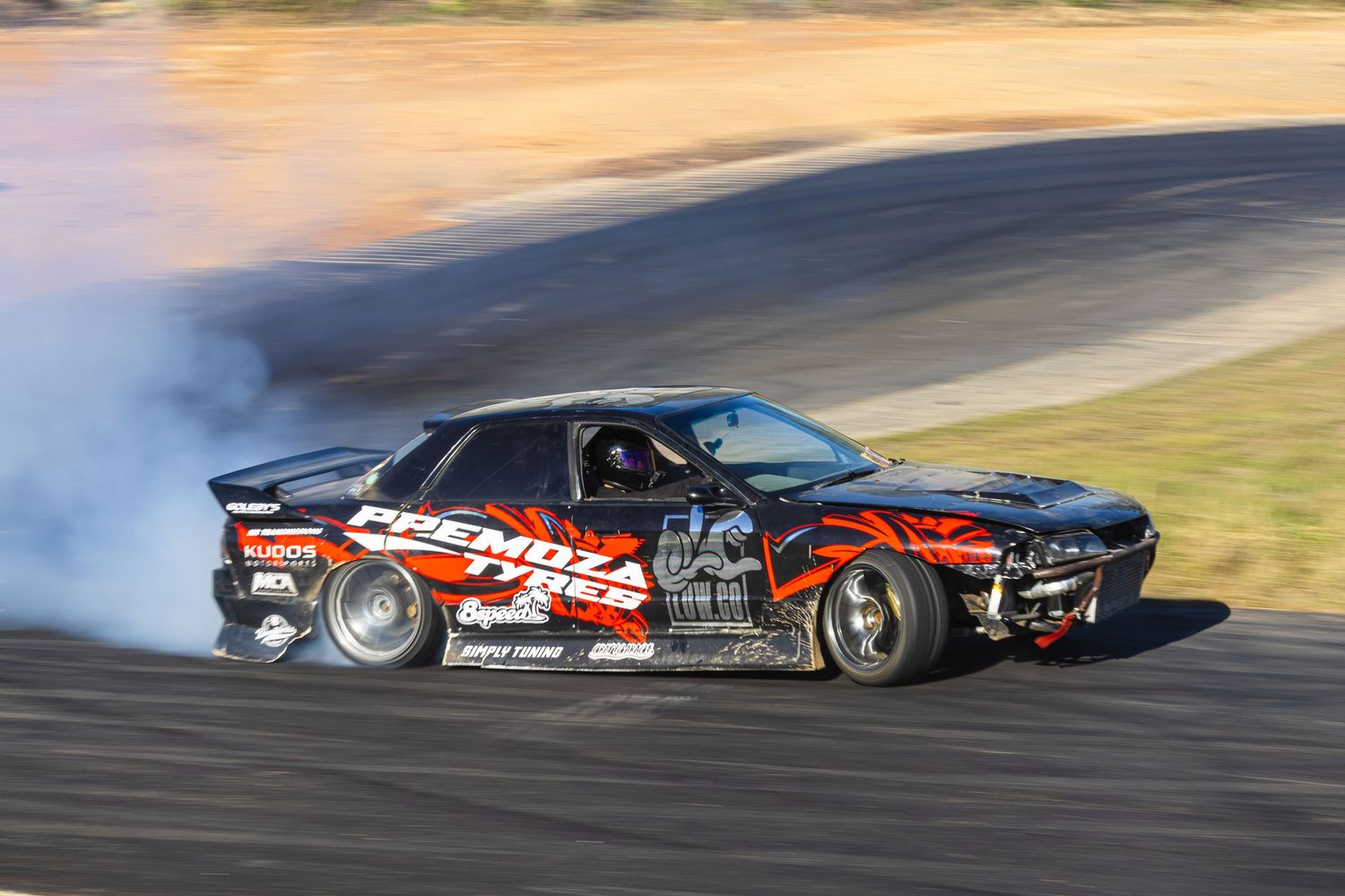 Black drift car with red flames livery producing thick tire smoke on racing circuit.