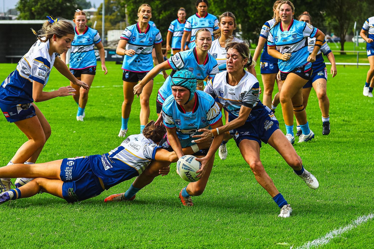 Women rugby league players in blue and white uniforms compete for the ball during an intense match on a green field.