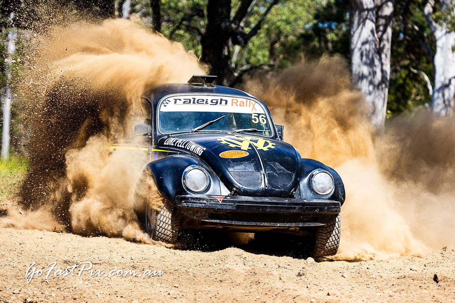 Black VW Beetle rally car kicking up massive dust cloud on dirt track during off-road race.