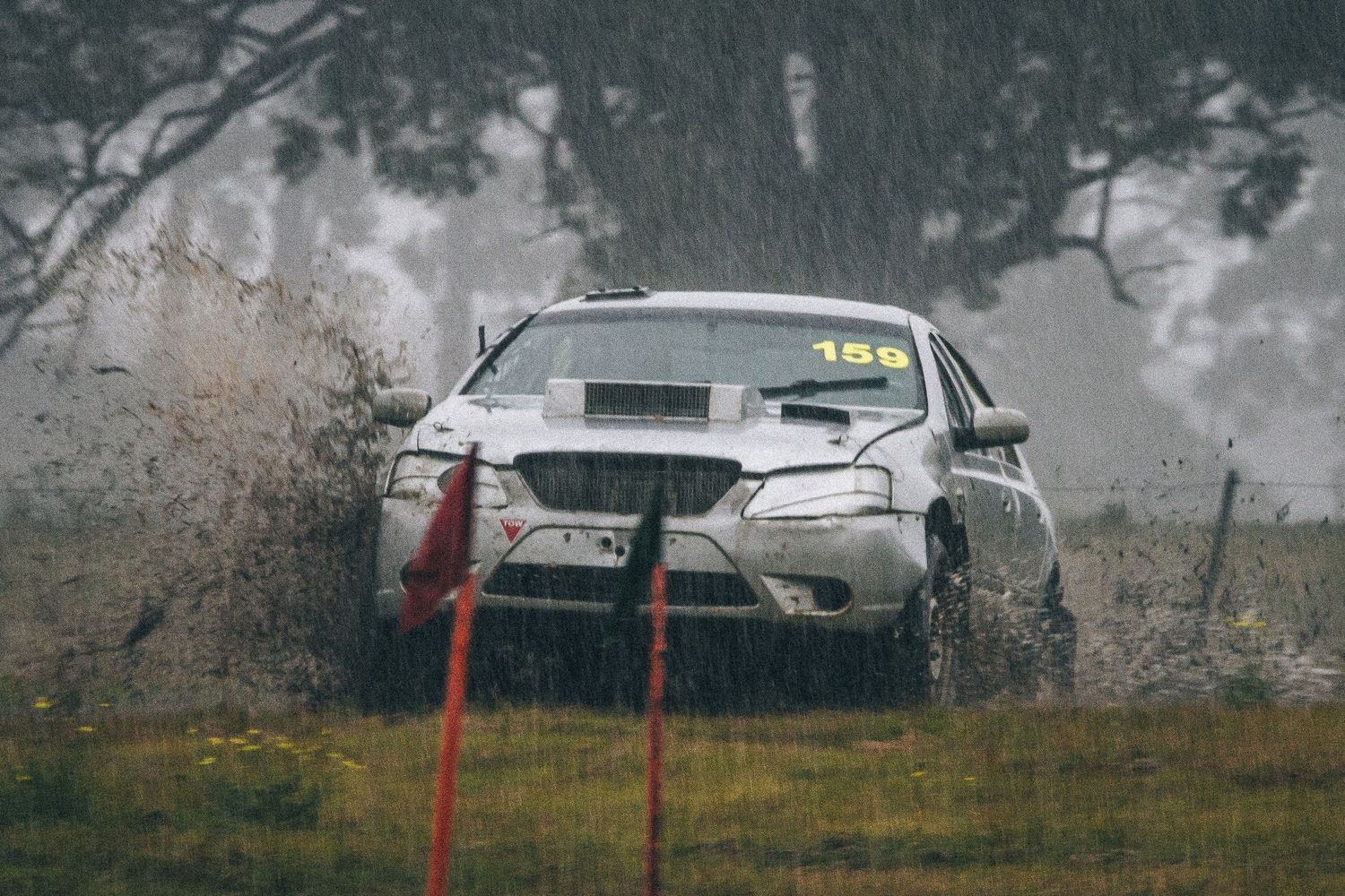 Silver rally car number 156 splashing through wet grass in heavy rain during motorsport event.