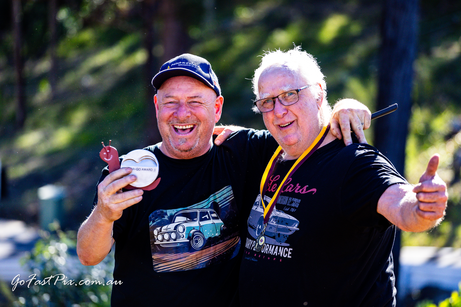 Two smiling men celebrating with a medal at an outdoor motorsport event.