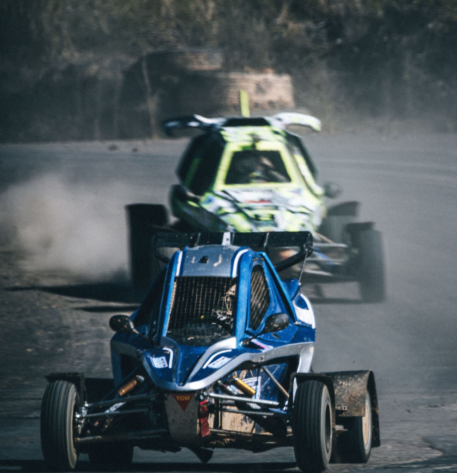 Blue open-frame racing buggy leads green competitor on dusty off-road motorsport track.