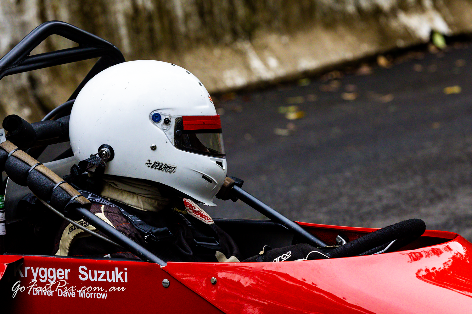 Racing driver wearing a white helmet seated in a red open-wheel formula race car on track.