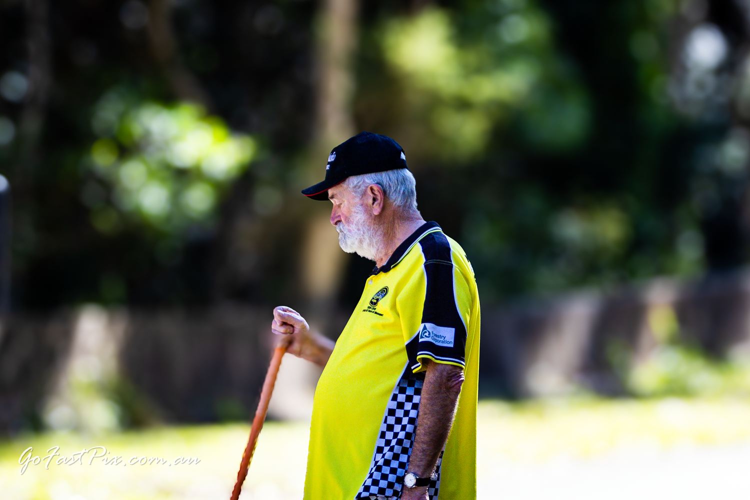 Elderly motorsport official in yellow vest walks with stick at racing event.