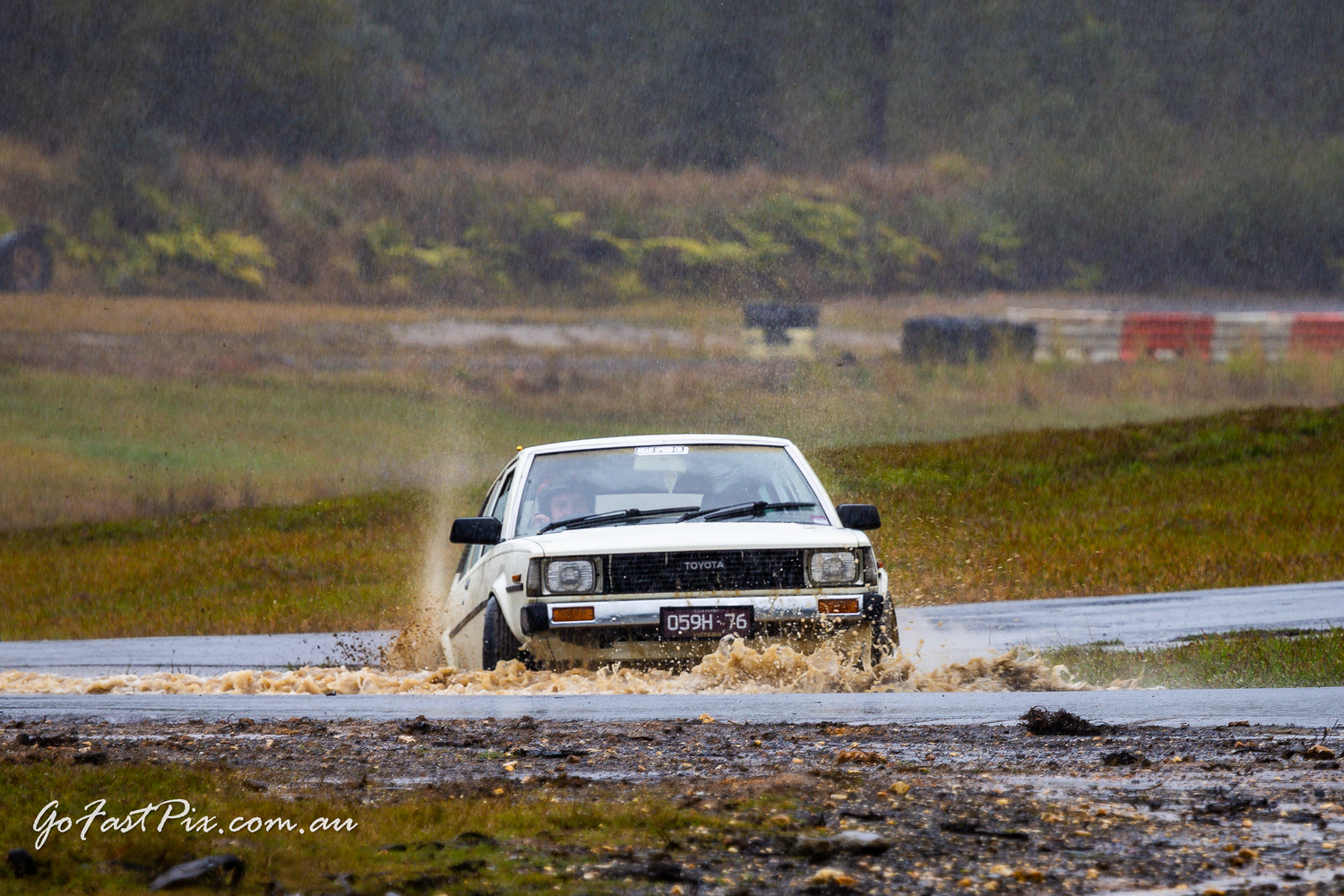 White rally car splashes through muddy water on wet rally stage.