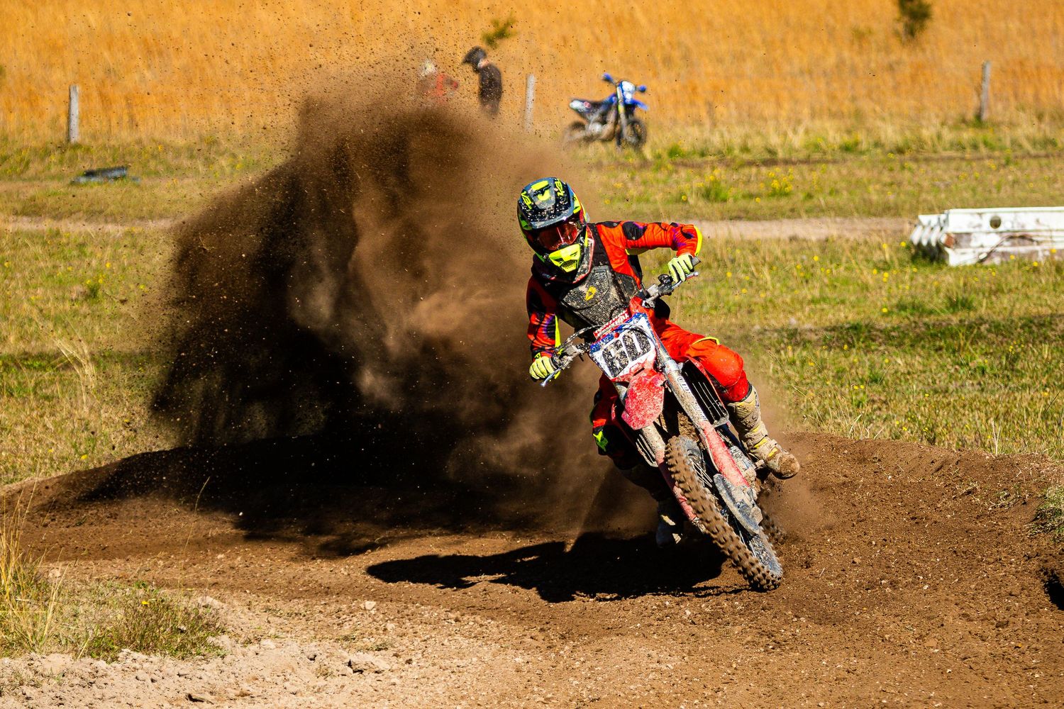 Motocross rider on red dirt bike kicking up dust cloud while racing on outdoor track in sunny conditions.