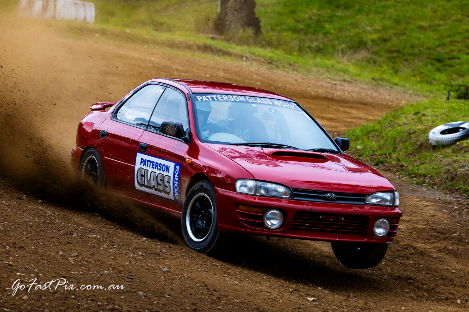 Red Subaru Impreza rally car racing on a dirt track, spraying dust during a competitive rally event.