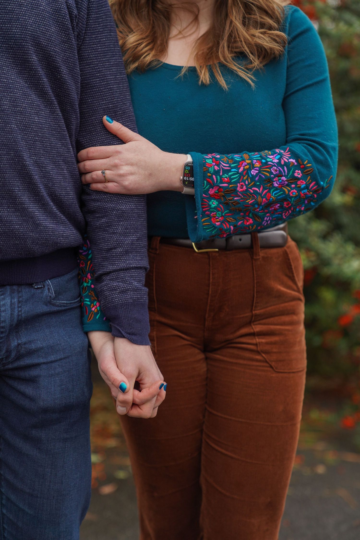 A couple holds hands and stands close together, the woman in a teal top and rust corduroy pants, during a fall engagement session.
