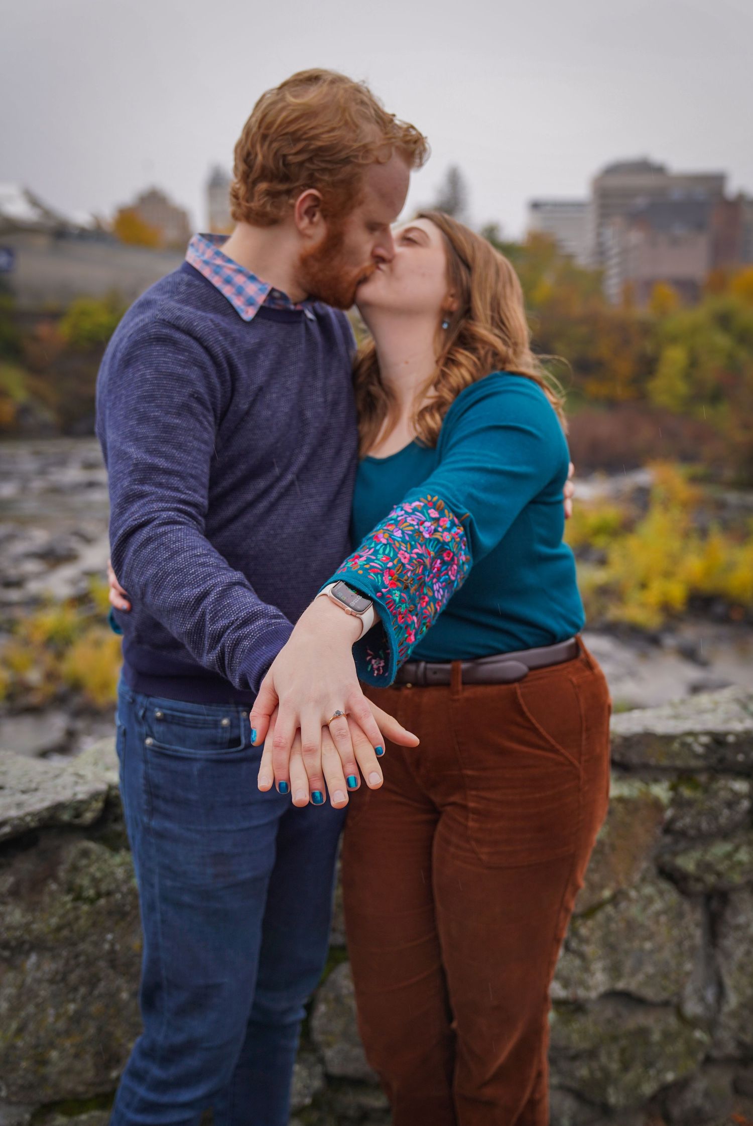 A man leans in to kiss his partner on the cheek as she smiles, surrounded by autumn foliage during an outdoor couples session.