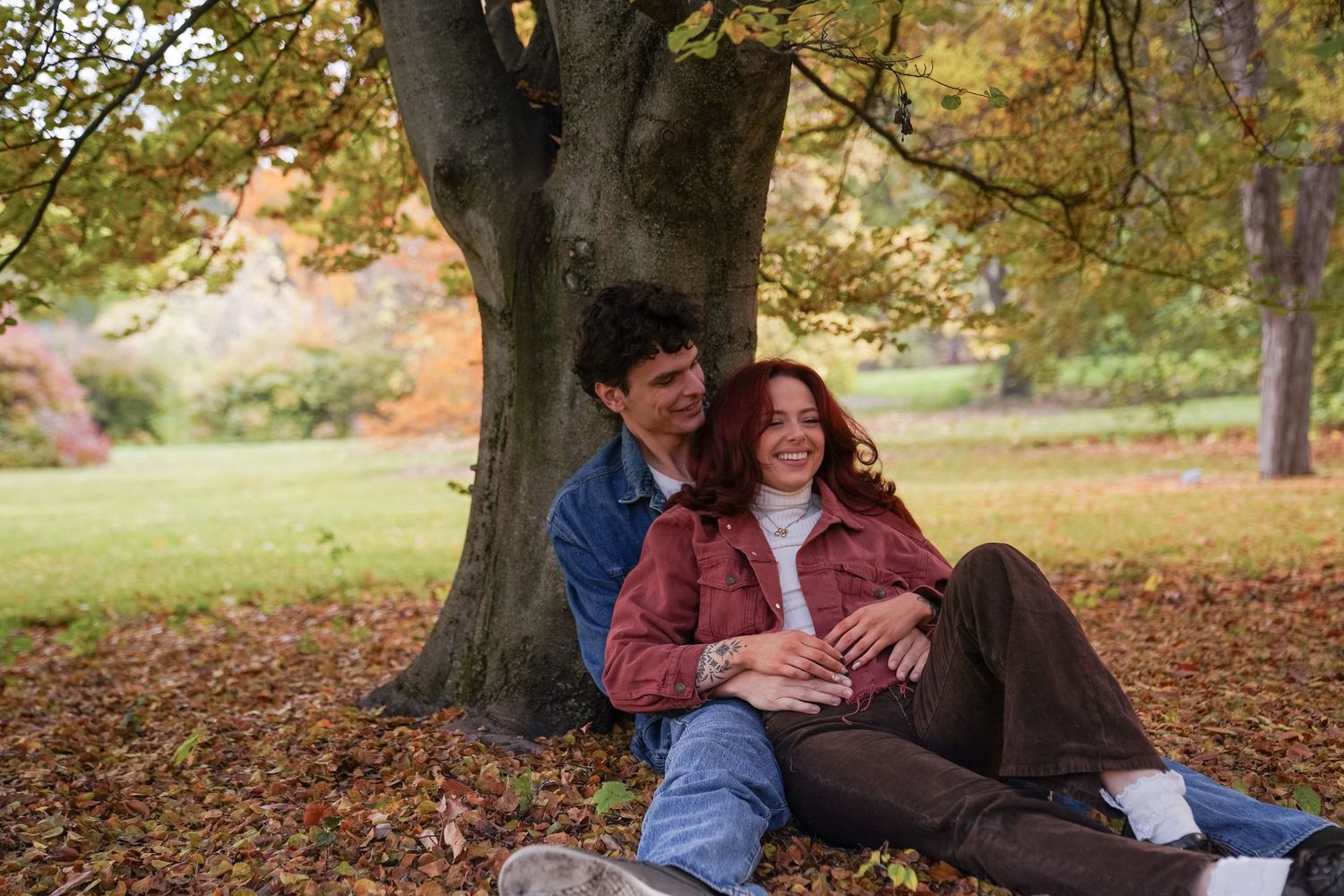 A couple sits relaxed together against the base of a large tree, laughing softly among fallen leaves during a fall couples photo session.