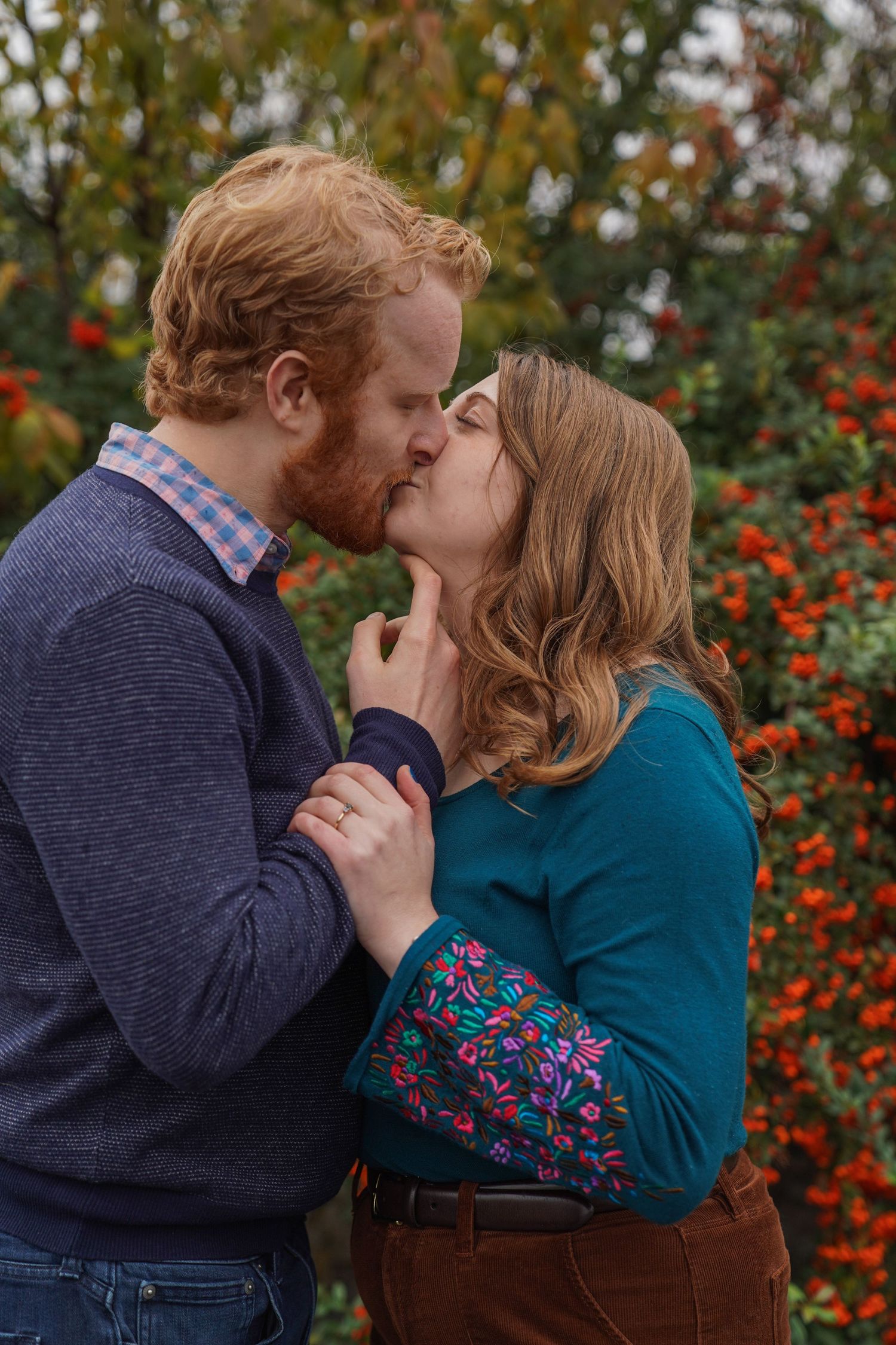 A couple shares a kiss surrounded by vibrant red autumn foliage during an outdoor fall engagement session.