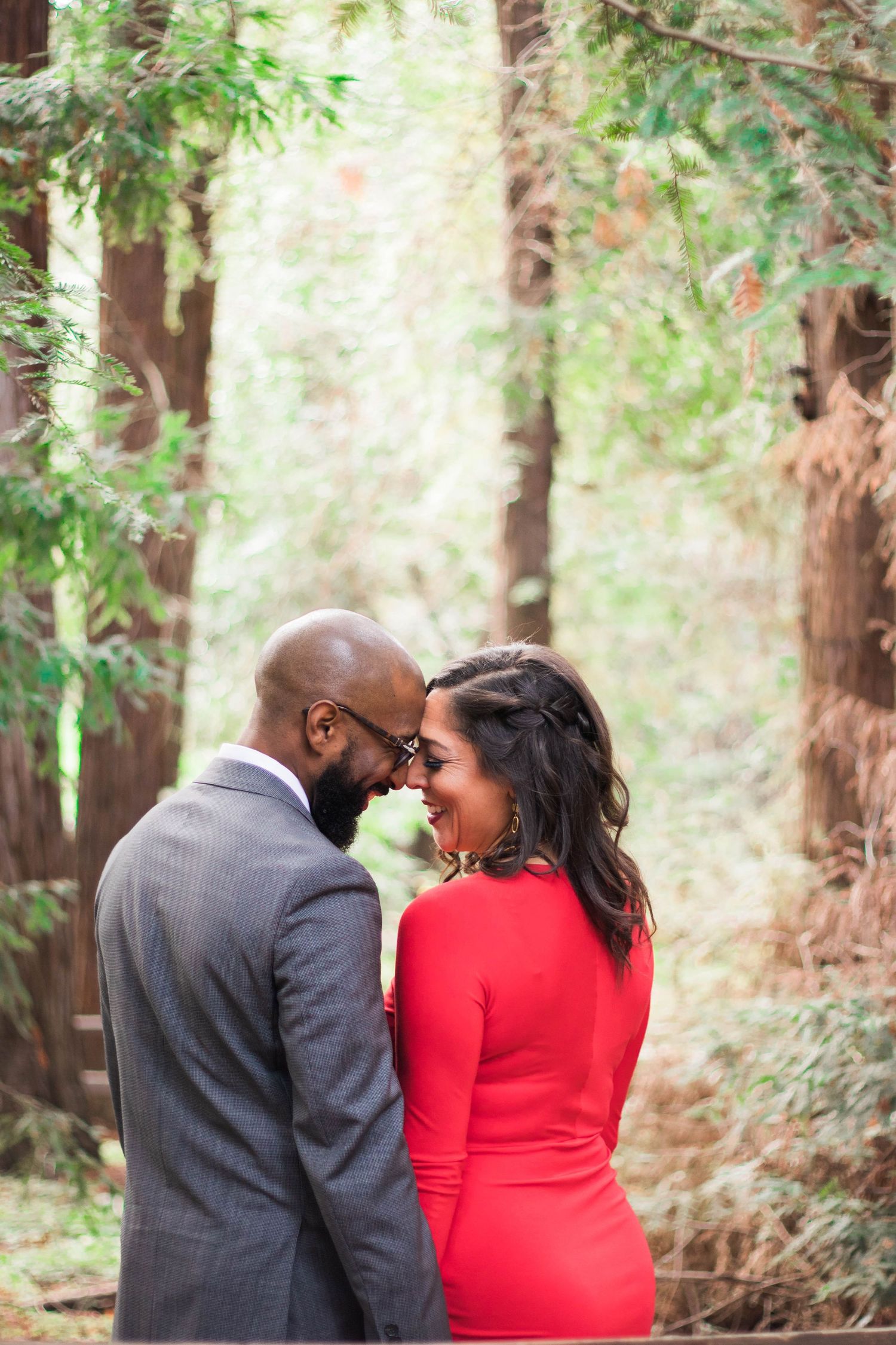 A couple stands close together among sun-lit trees, the woman in a red dress, sharing a tender moment during an outdoor engagement session.