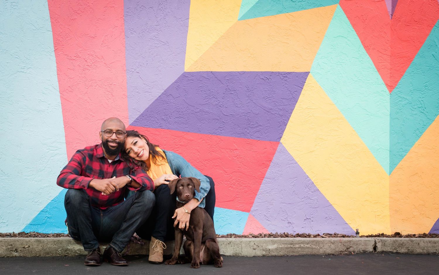 A couple sits casually on the ground in front of a bright geometric mural, their dog resting beside them, smiling warmly at the camera.
