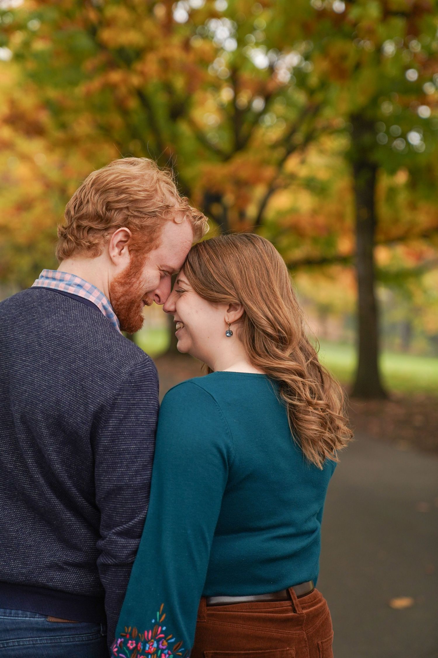 A couple stands nose-to-nose beneath golden autumn trees, sharing a quiet, tender moment during a fall engagement session.