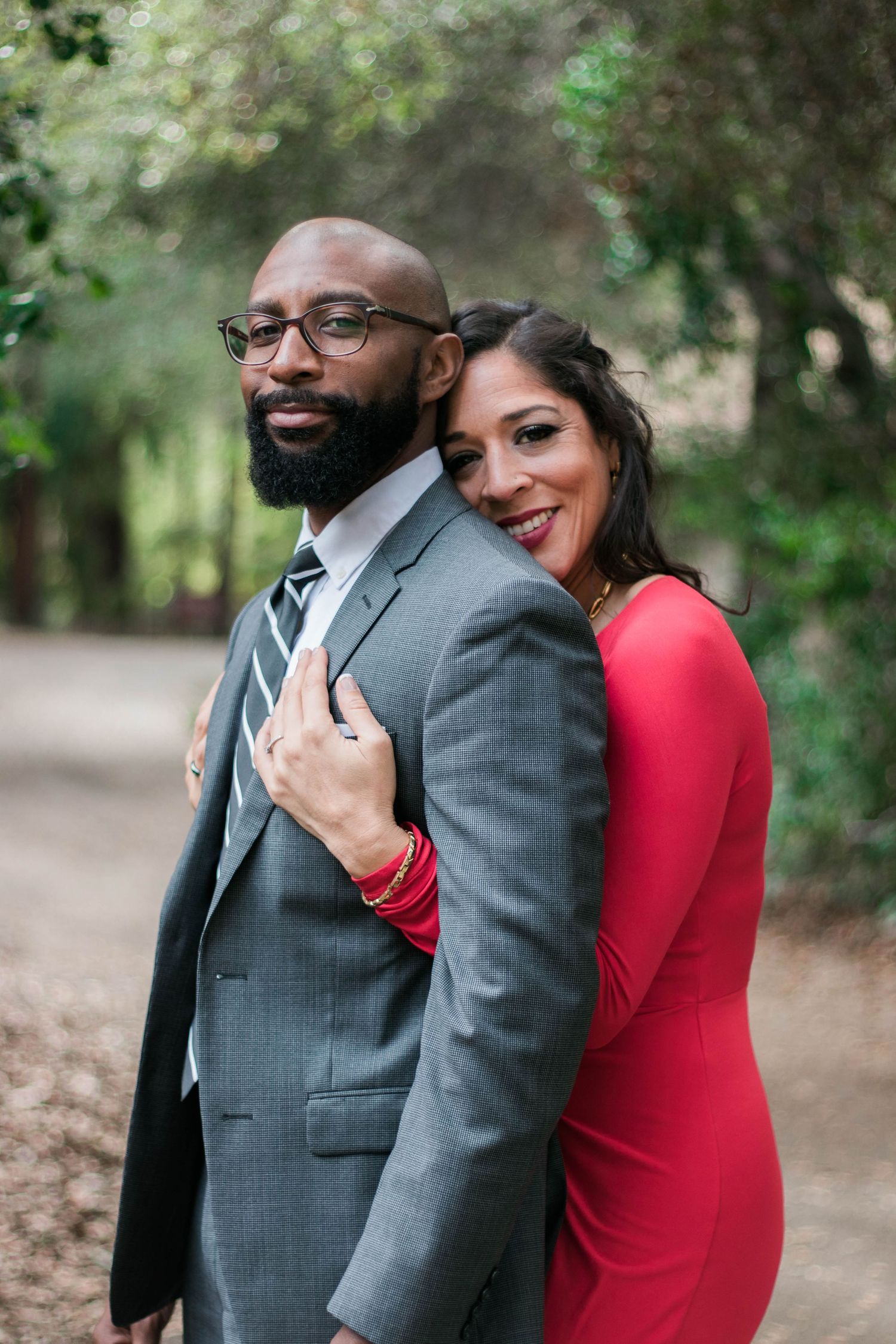 A couple poses together on a wooded path, the woman in a red dress leaning into her partner during an outdoor engagement session.