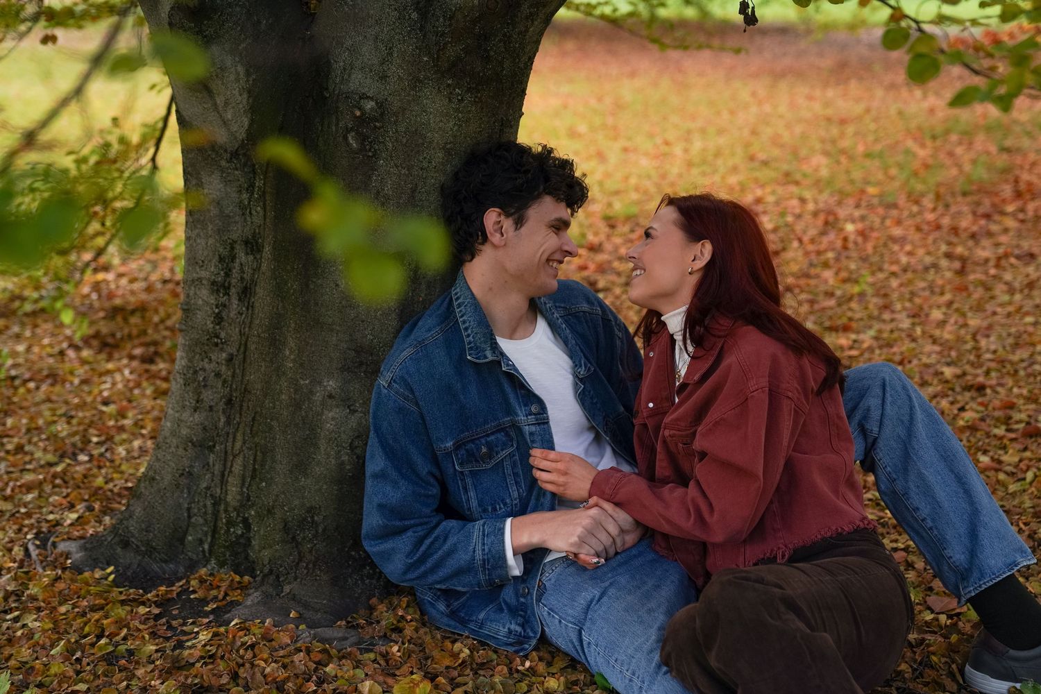 A couple sits close together at the foot of a tree, smiling and talking naturally among autumn leaves during their couples photo session.
