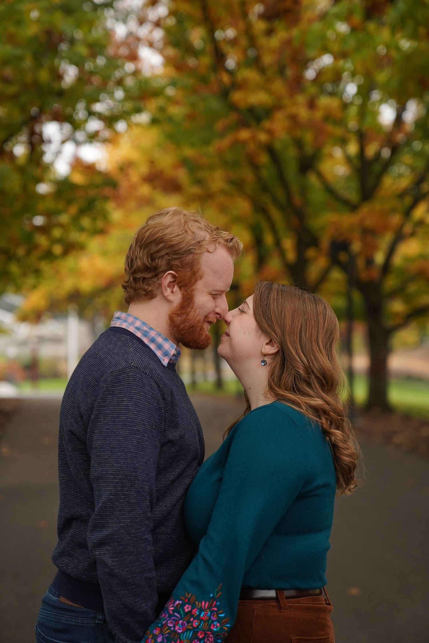 A couple leans close together on a tree-lined path in peak fall color, captured during an intimate outdoor engagement session.