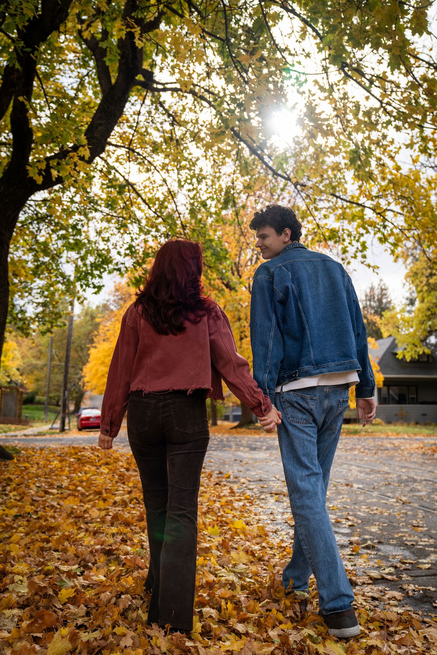 A couple walks hand in hand away from the camera down a sun-lit autumn path, golden leaves falling around them.