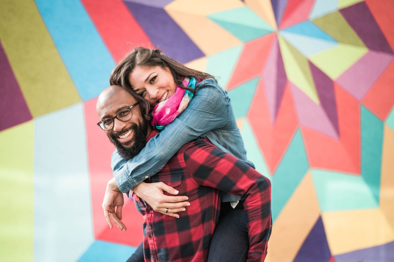 A woman hugs a man from behind, both laughing, posed in front of a vibrant multicolored mural during an engagement session.