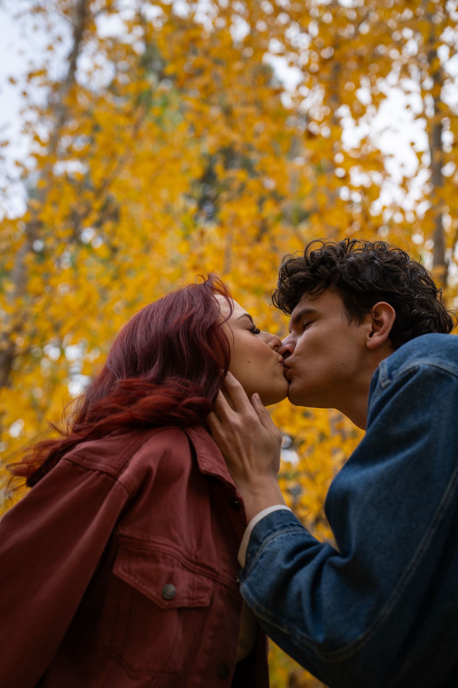 A couple shares a gentle kiss against a backdrop of golden autumn foliage during an outdoor fall couples photo session.