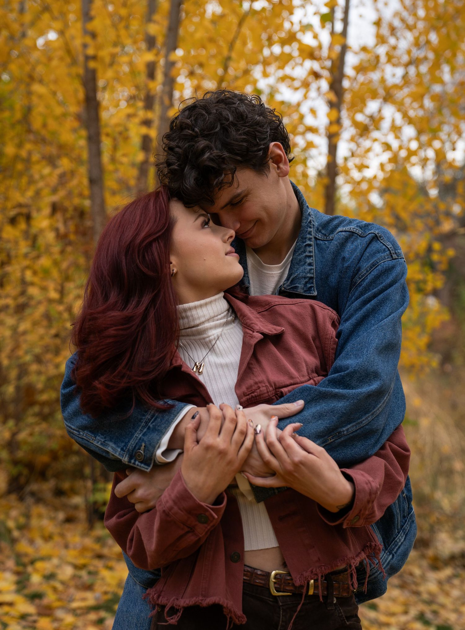 A couple embraces and shares a kiss among golden fall foliage, the woman's auburn hair vivid against the warm autumn colors.