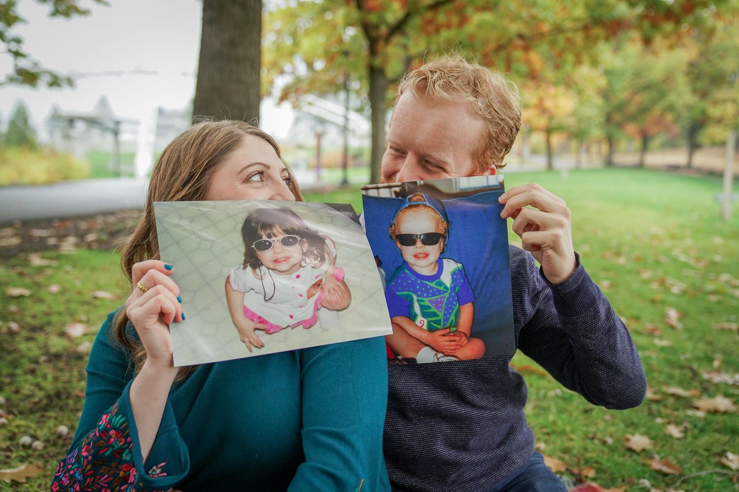 A couple holds up playful printed photos of themselves in a park, laughing together during a fun fall engagement session.