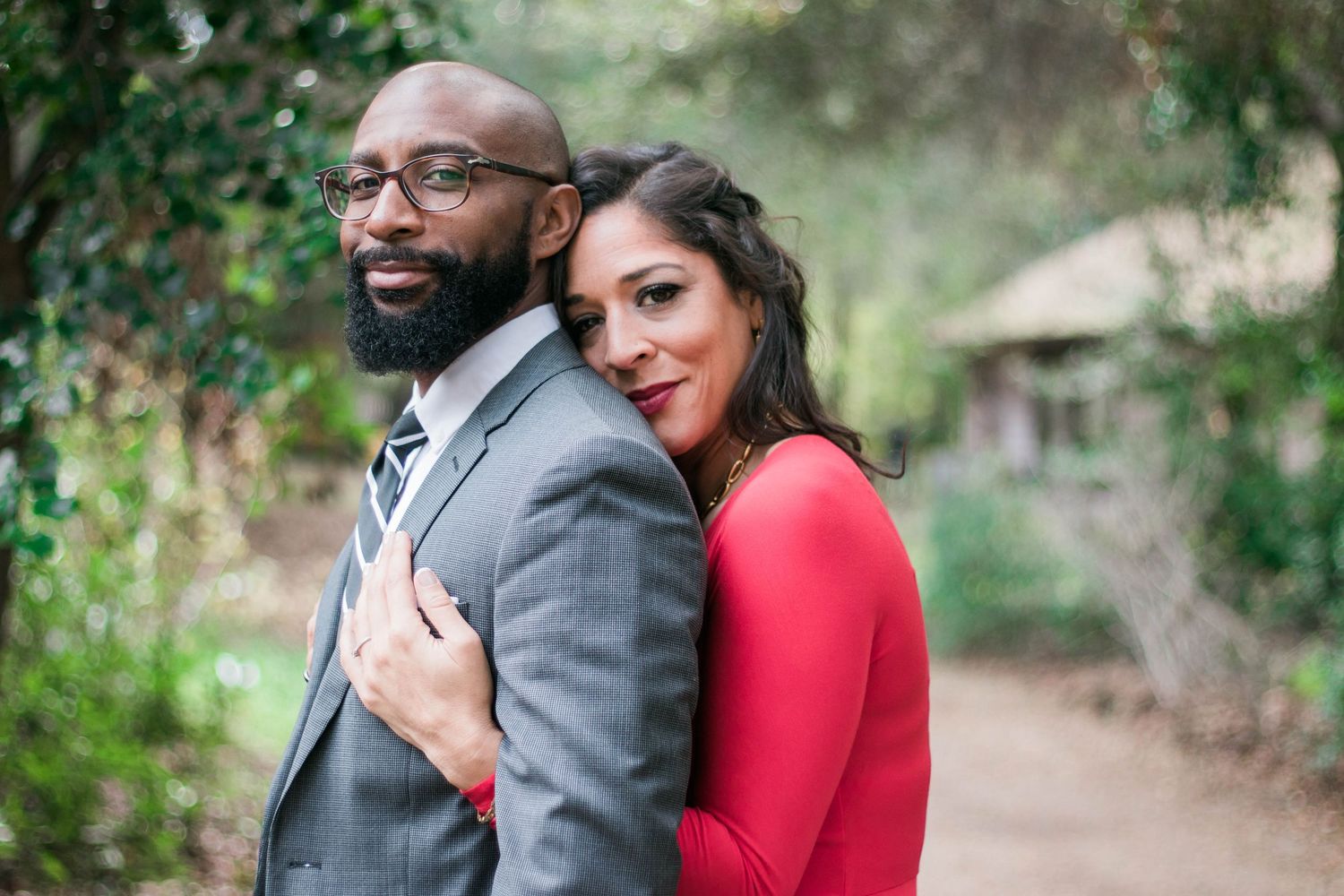 A woman in a red dress rests her hands on her partner's shoulders as they pose together on a lush garden path for their engagement photos.