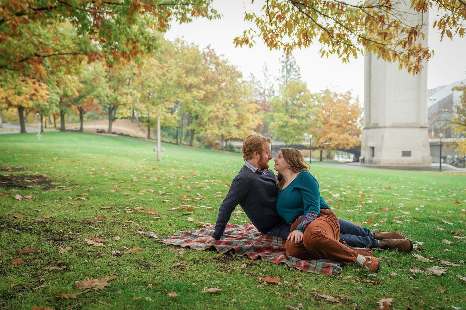 A couple sits together on a blanket of fallen leaves in a park, relaxed and laughing during an autumn engagement session.
